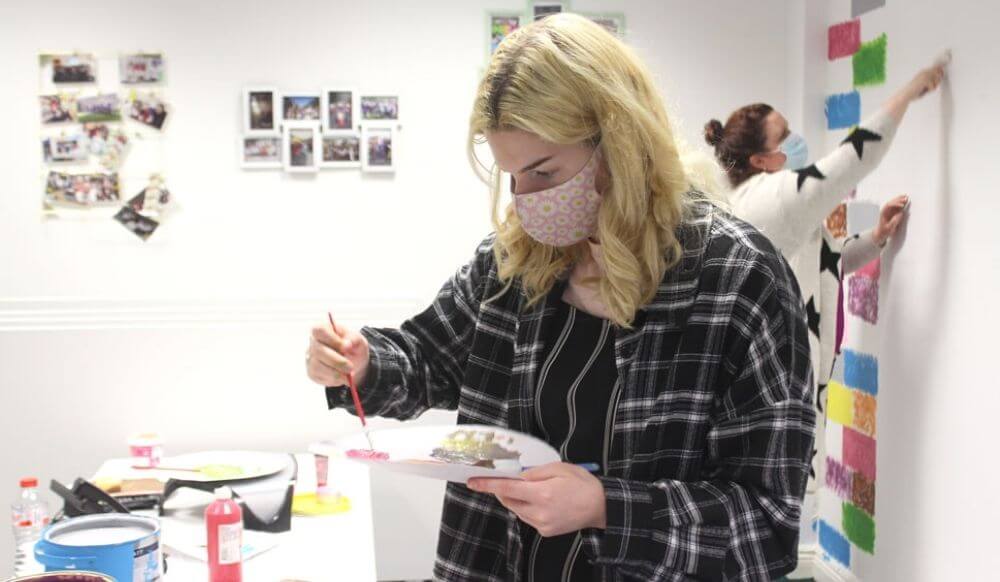 A young woman mixes paint in the foreground while another woman paints in the background