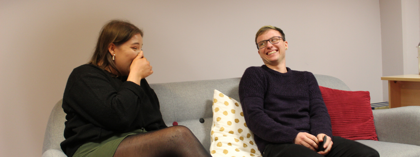 Two young people, girl and boy sitting down laughing on a grey couch