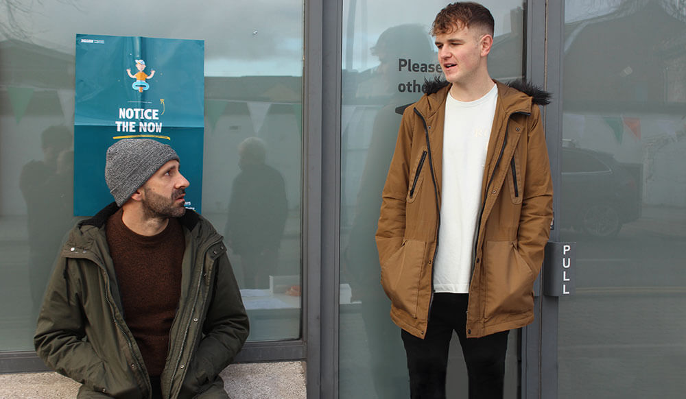 A young man standing talking to an older man who is sitting down