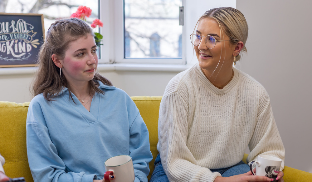 Two girls are sitting down on a yellow sofa, talking. There are mugs in their hands.